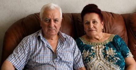 An elderly couple sitting together at a table with an old metal box containing decades of hidden savings and family documents.